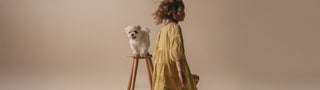 Girl in yellow dress next to small white dog on stool on beige background