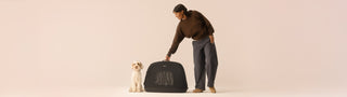 Woman smiles down at a fluffy white dog seated next to the Crispin pet travel crate against a beige background.