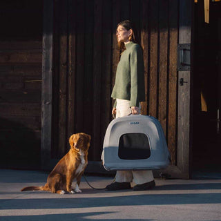 Woman holding a Crispin pet travel crate with a dog sitting on the ground in front of a wooden building.