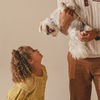 Smiling child looks up at small white dog held by adult.